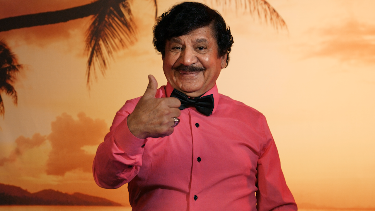 Karim visar tummen upp mot en bakgrund med palmer och solnedgång.