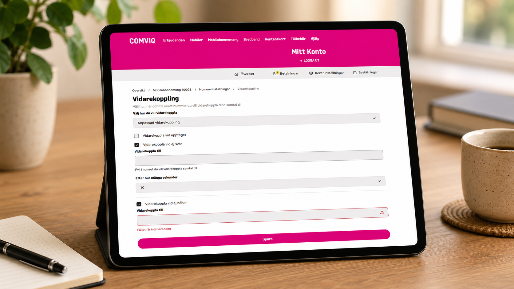 Bildbeskrivning: En surfplatta står på ett träbord i en lugn inomhusmiljö. Surfplattan är i fokus och på skärmen visas Comviqs sida för Vidarekoppling i Mitt Konto. Sidan innehåller inställningar för att vidarekoppla samtal vid upptaget, vid uteblivet svar eller när telefonen inte är nåbar. Formulärfält för telefonnummer och val av antal sekunder innan vidarekoppling visas, tillsammans med en rosa Spara-knapp längst ned. Bakgrunden är mjukt suddig och innehåller bland annat en kaffekopp, en anteckningsbok och en grön växt.