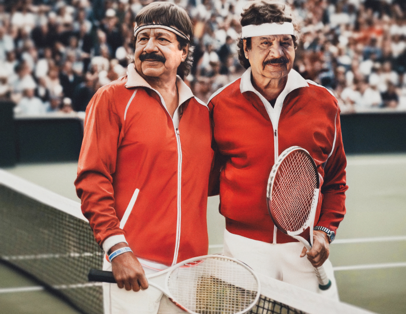 Två äldre män, väldigt lika varandra, med mustasch i röda träningsjackor och pannband håller tennisracketar på en tennisbana med publik.