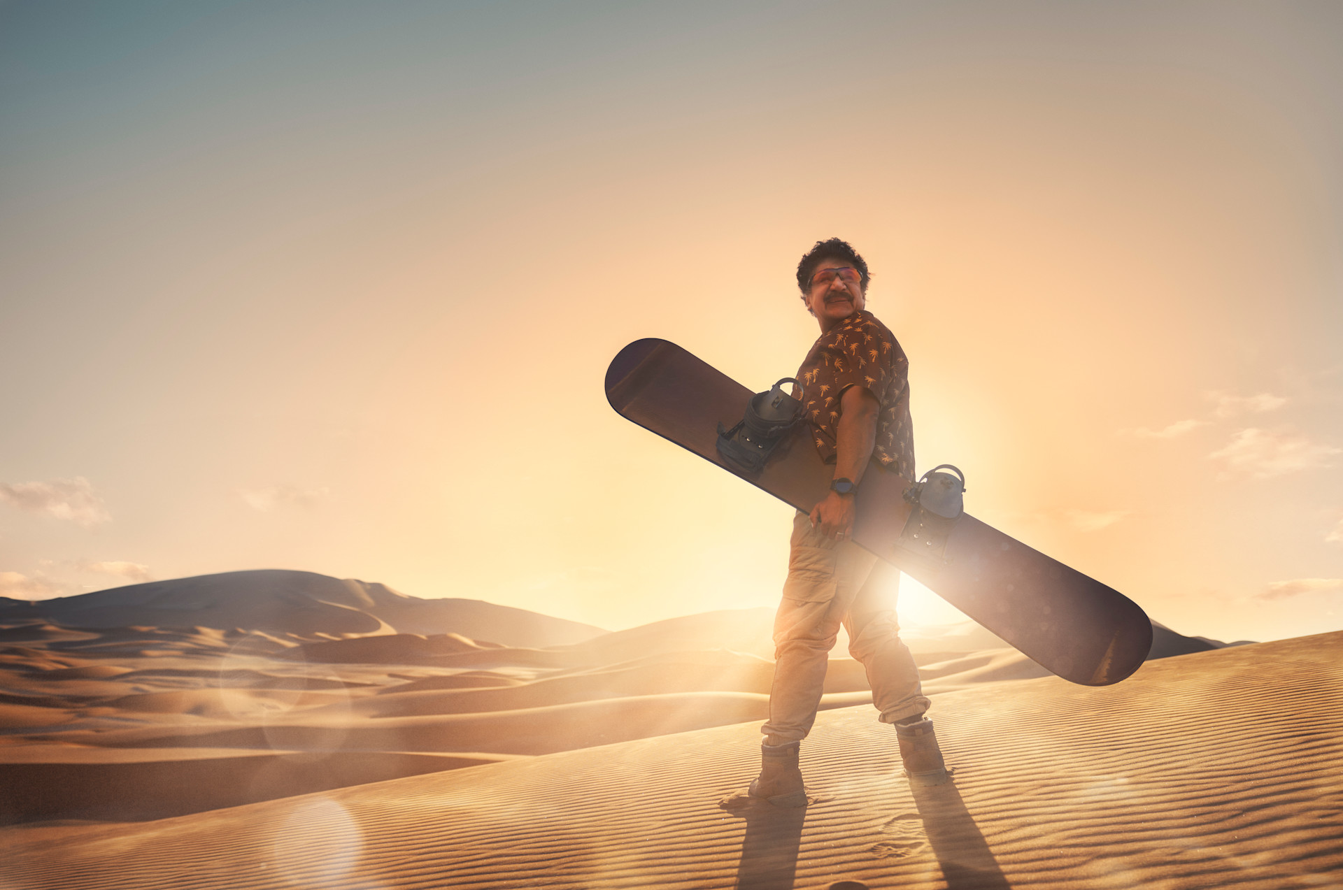 Person med snowboard står i ökenlandskap vid solnedgång, gyllene ljus lyser över sanddynerna.