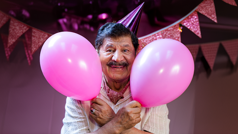 En leende äldre man med partyhatt håller två rosa ballonger omgivna av rosa vimplar i bakgrunden.