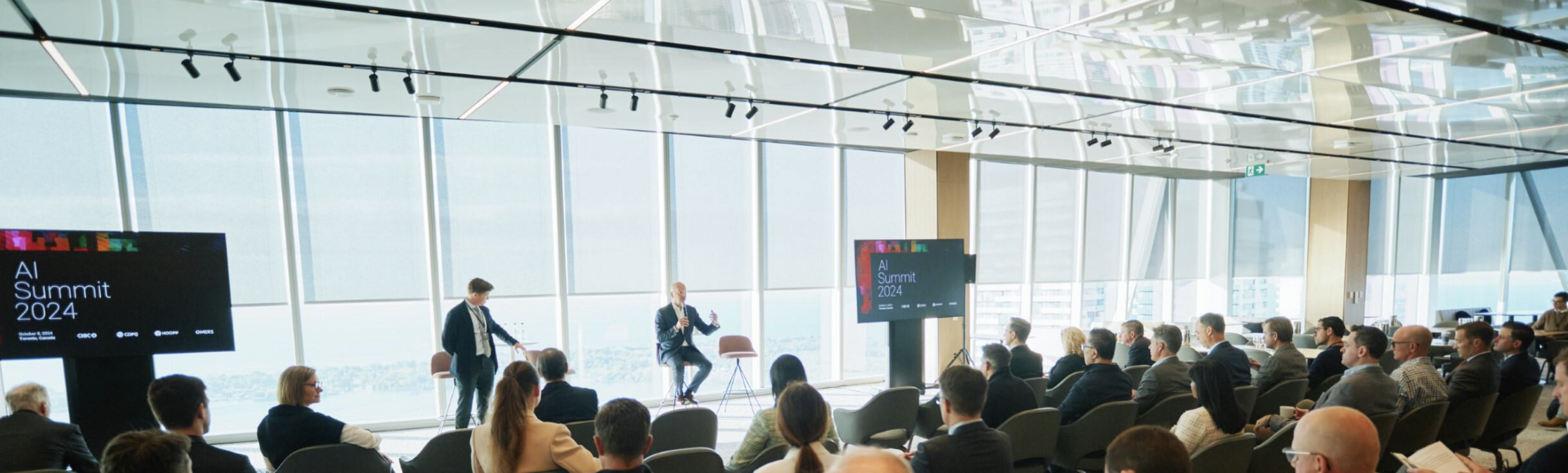 Two men sitting and speaking in front of a crowd at an AI summit