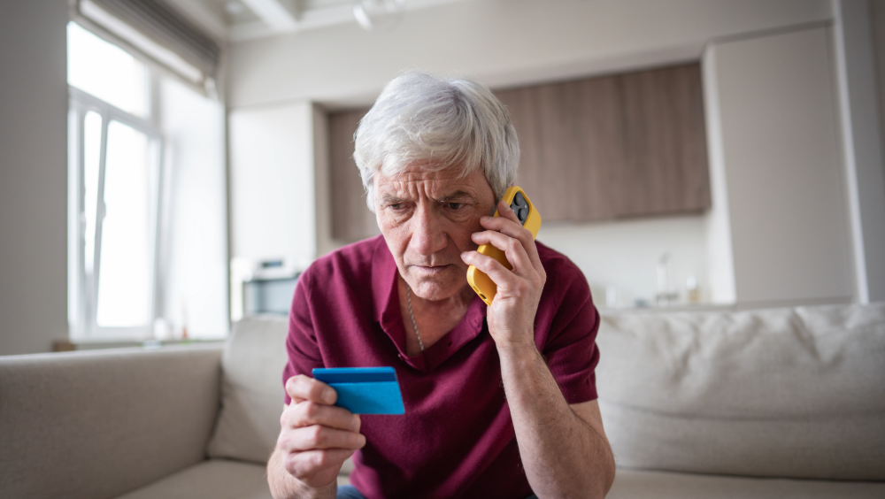 An elderly man sitting on a couch holding a cell phone and a credit card