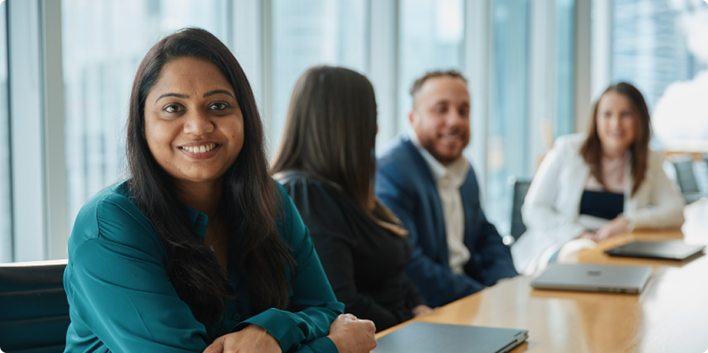 Woman sitting at a boardroom table smiling at the camera while three coworkers chat beside her