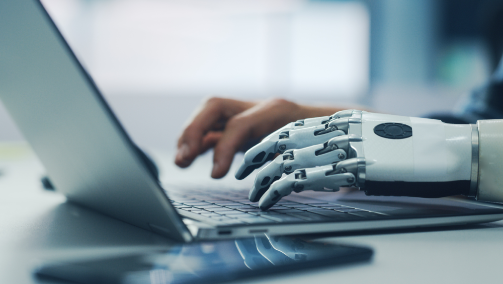 A person with a robot hand typing on a laptop