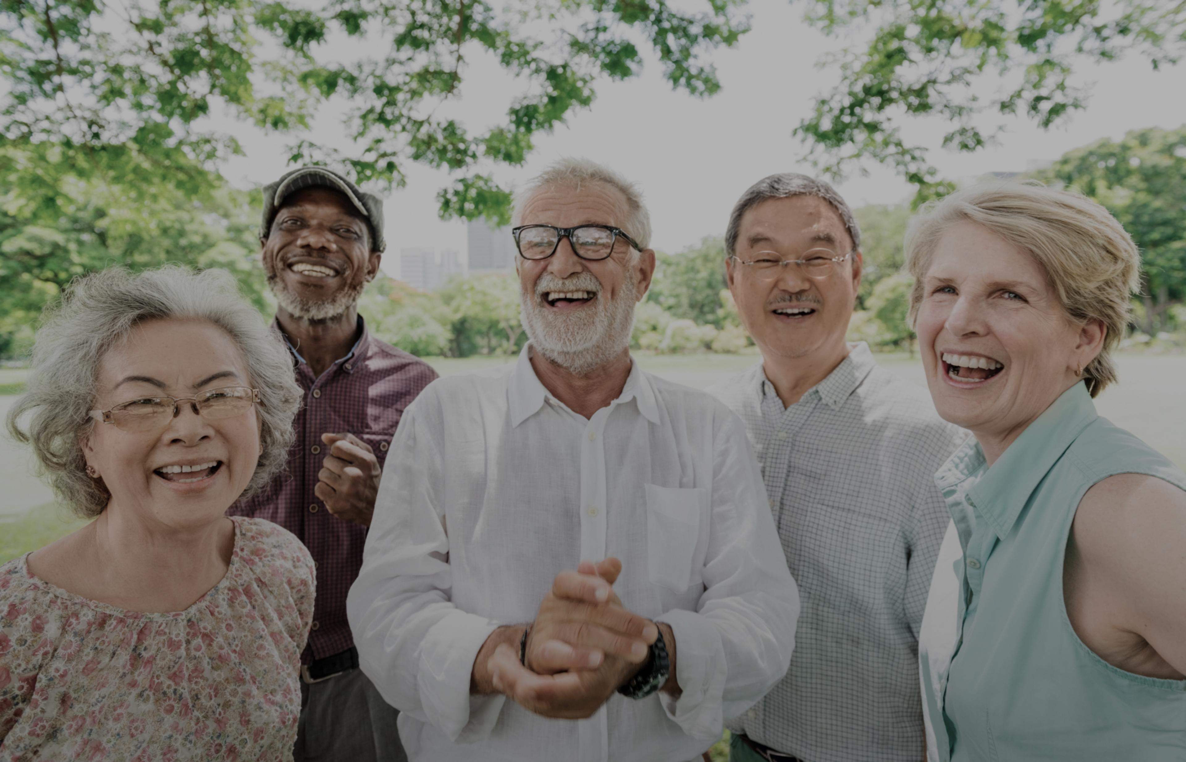 A group of elderly people standing together laughing and smiling for the camera with a field in the background and a tree over head.