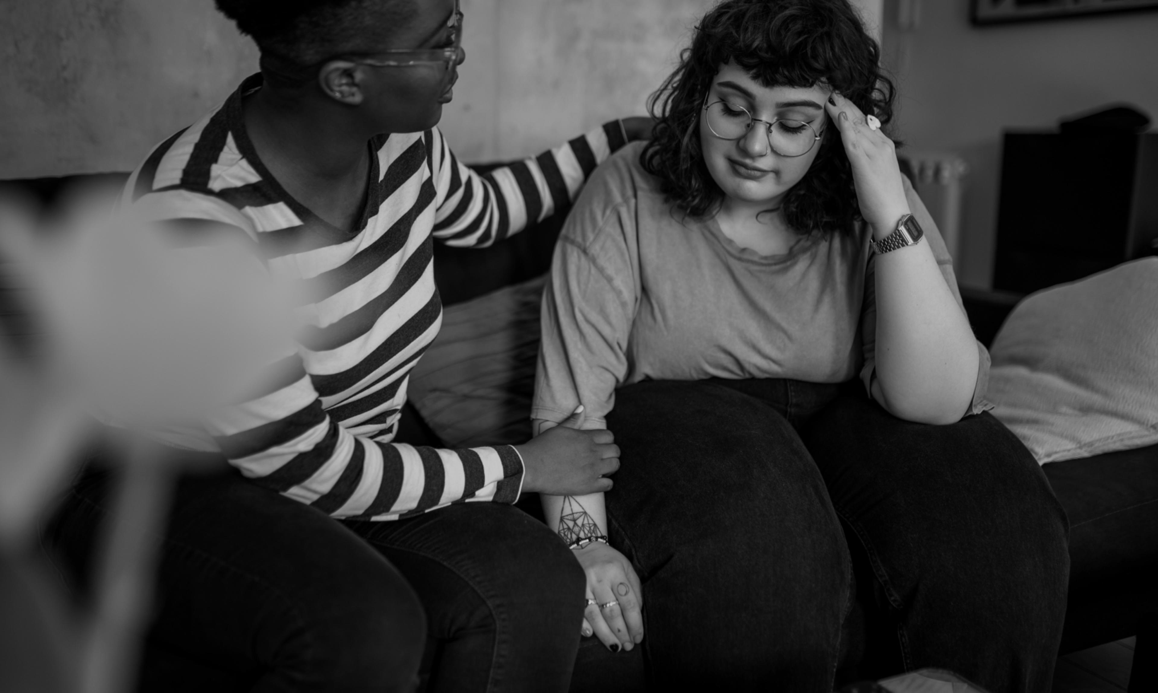 Two young women sitting on a couch while one is comforting the other who seems to be distressed.