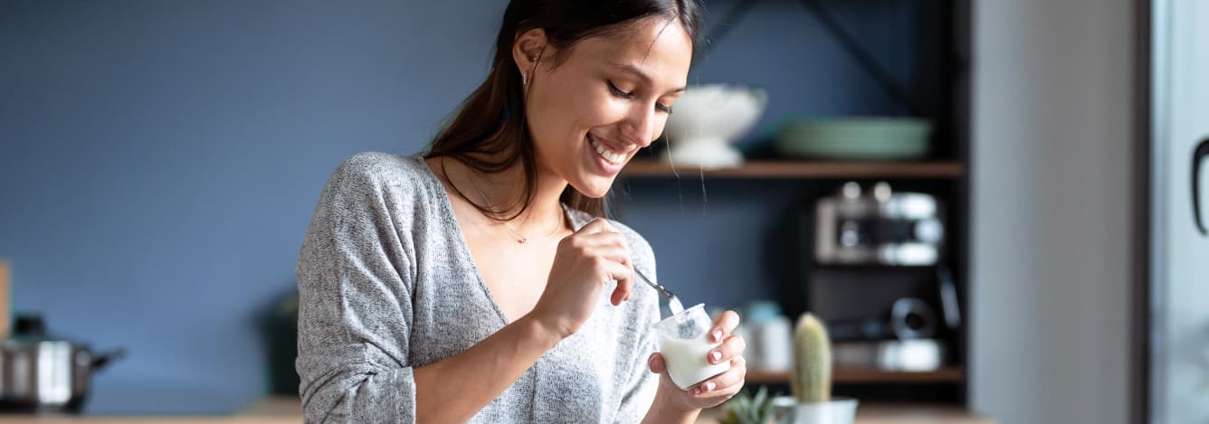 A smiling young woman in a grey sweater eating yogurt with a spoon in a modern kitchen.