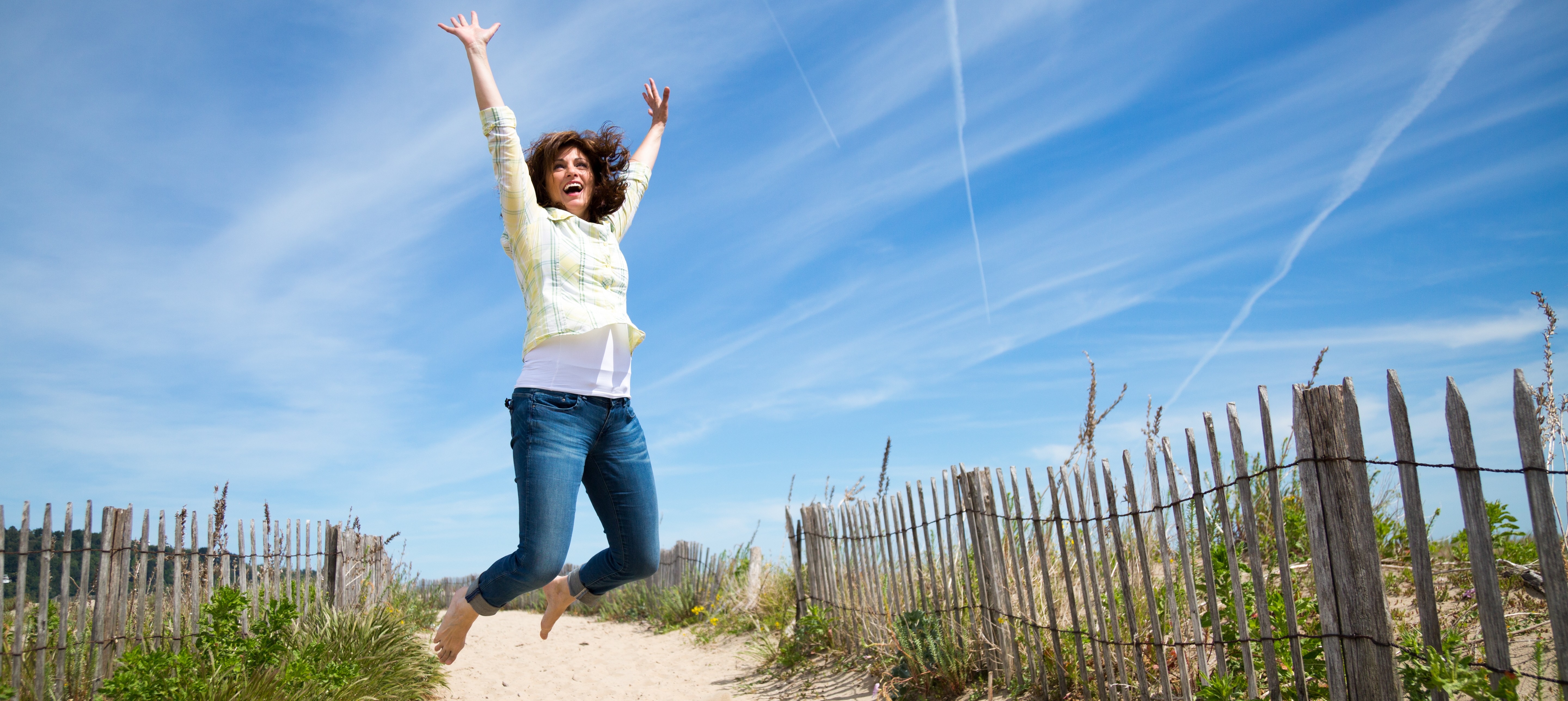 A joyful woman jumping with arms raised on a sandy beach path, wearing jeans and a yellow shirt, under a bright blue sky.