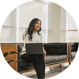 Kobieta o długich czarnych włosach siedzi na ławce z laptopem na kolanach i słucha uważnie współpracownika.