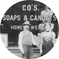 Czarno-białe zdjęcie mężczyzny i kobiety stojących przed witryną sklepową, na której widnieją słowa "CO'S. SOAPS & CANDLES, HONEST WEIGHT." Ikona jest otoczona niebieskim okręgiem.