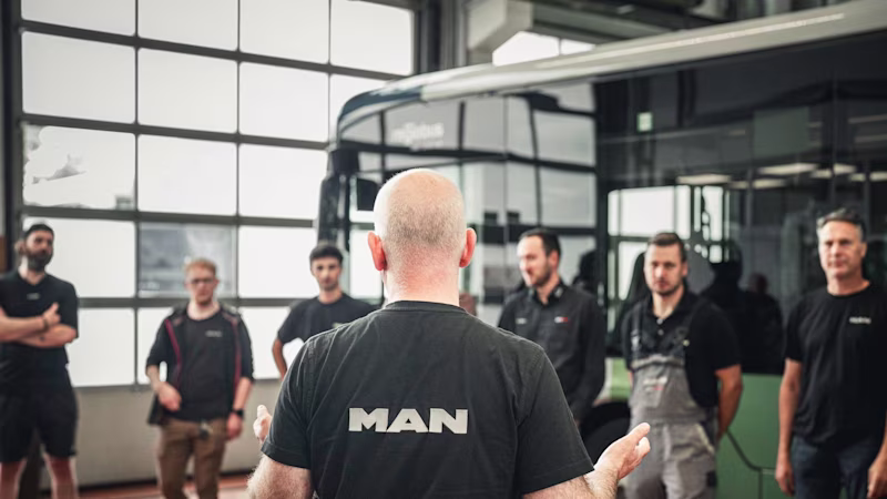 A foreman in a MAN T-shirt stands in front of a group of mechanics in the workshop in front of a bus.