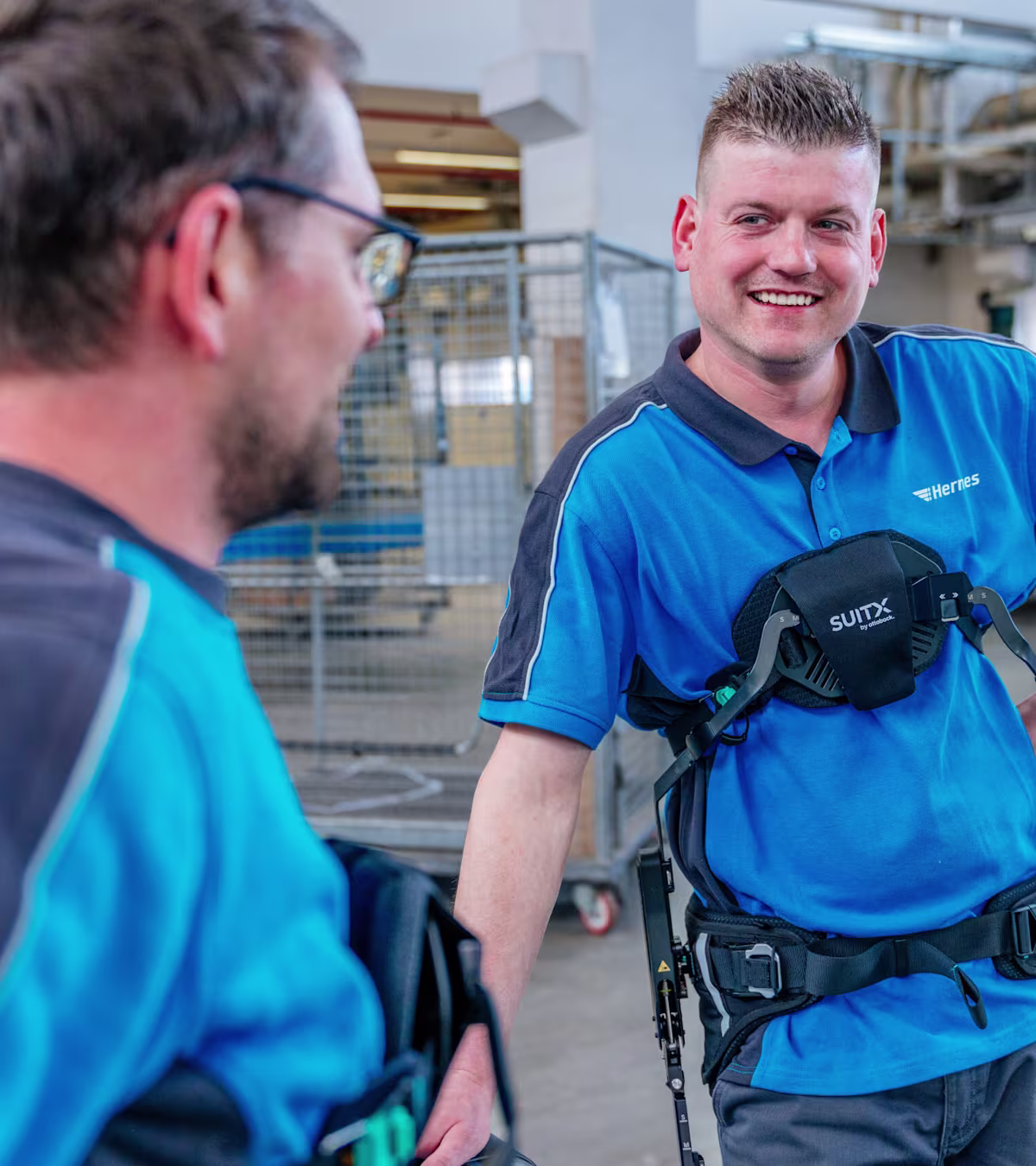 Zwei Lagerarbeiter mit Rückenexoskelett stehen an einem Förderband und lächeln.