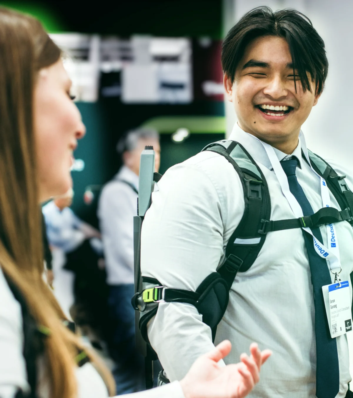 Male person wearing a SUITX exoskeleton at a trade show and smiling while talking to a woman in the foreground