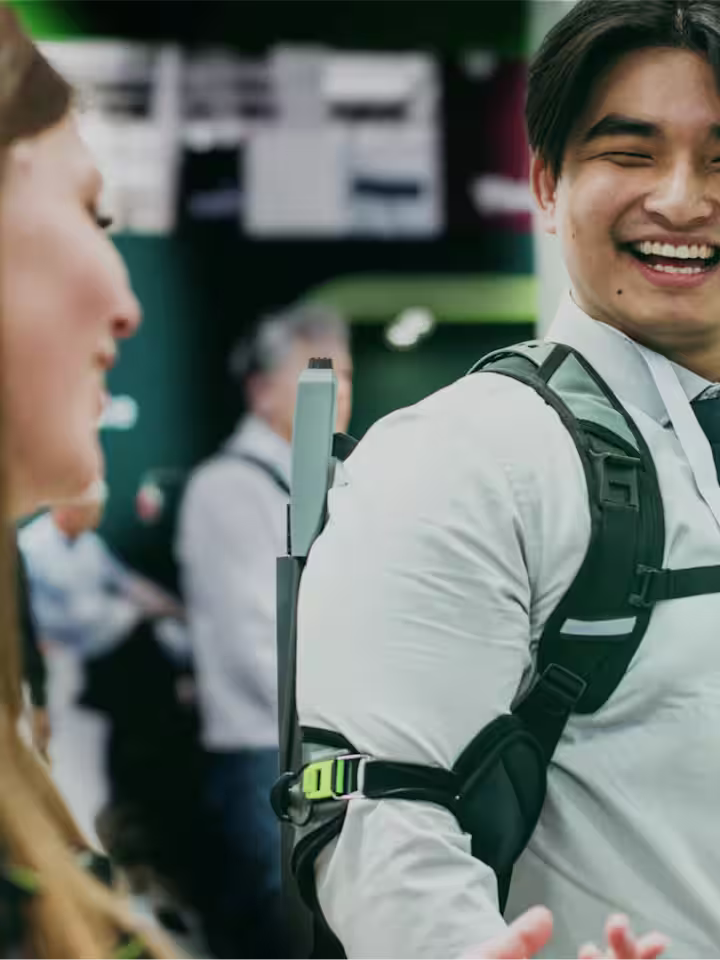 Male person wearing a SUITX exoskeleton at a trade show and smiling while talking to a woman in the foreground