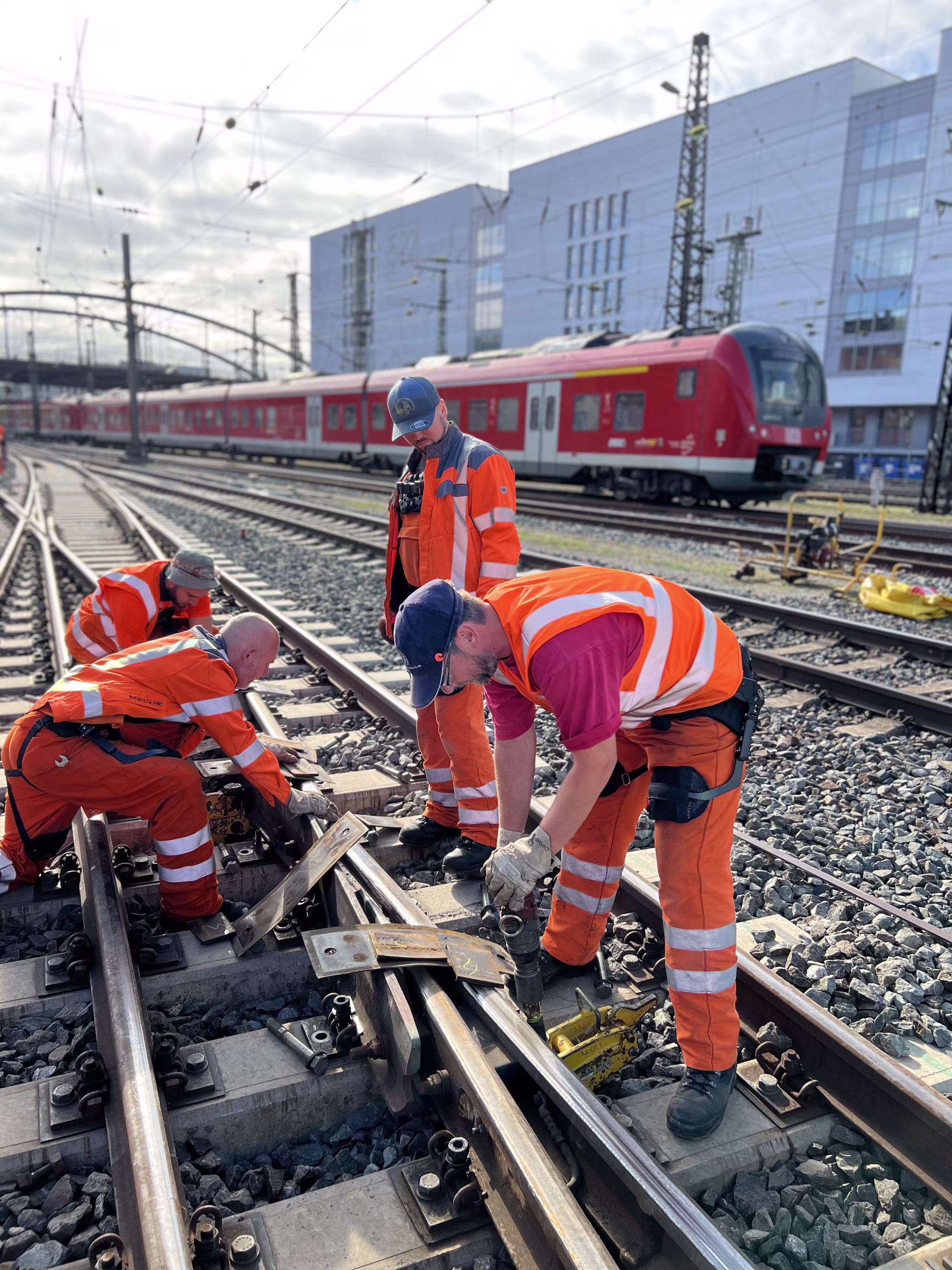 Four workers are working on a rail track. A train passes by in the background.