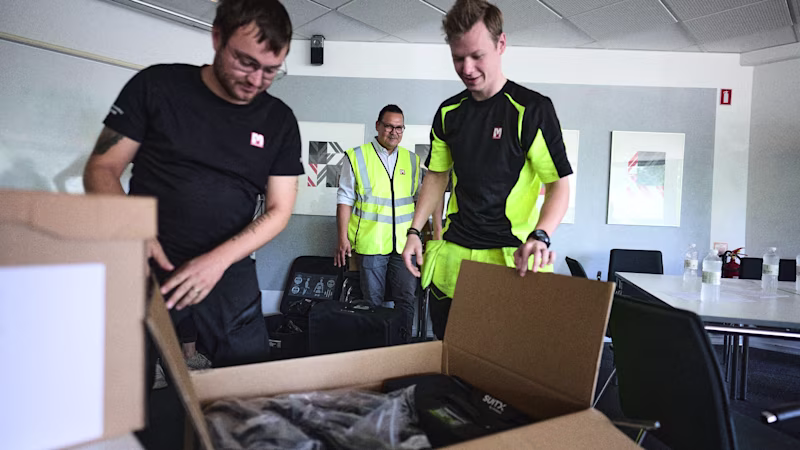 Zwei Mitarbeiter in Arbeitskleidung in einem Besprechungsraum packen einen Karton von SUITX aus.