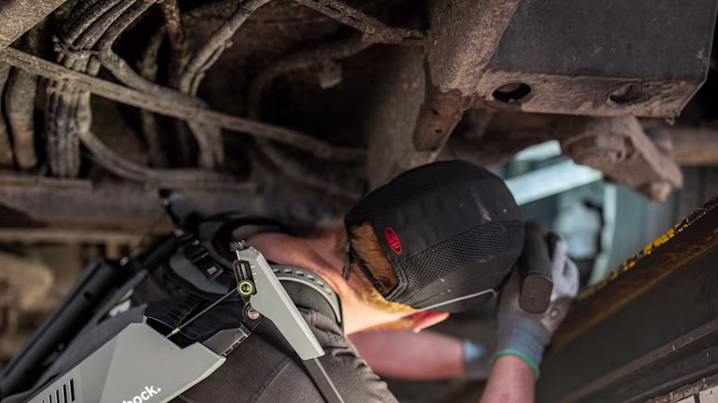 A mechanic stands in a ditch under a vehicle and repairs it. He is wearing a shoulder exoskeleton for support.