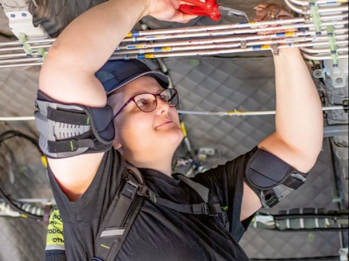 A worker lays cables overhead and wears a shoulder exoskeleton.