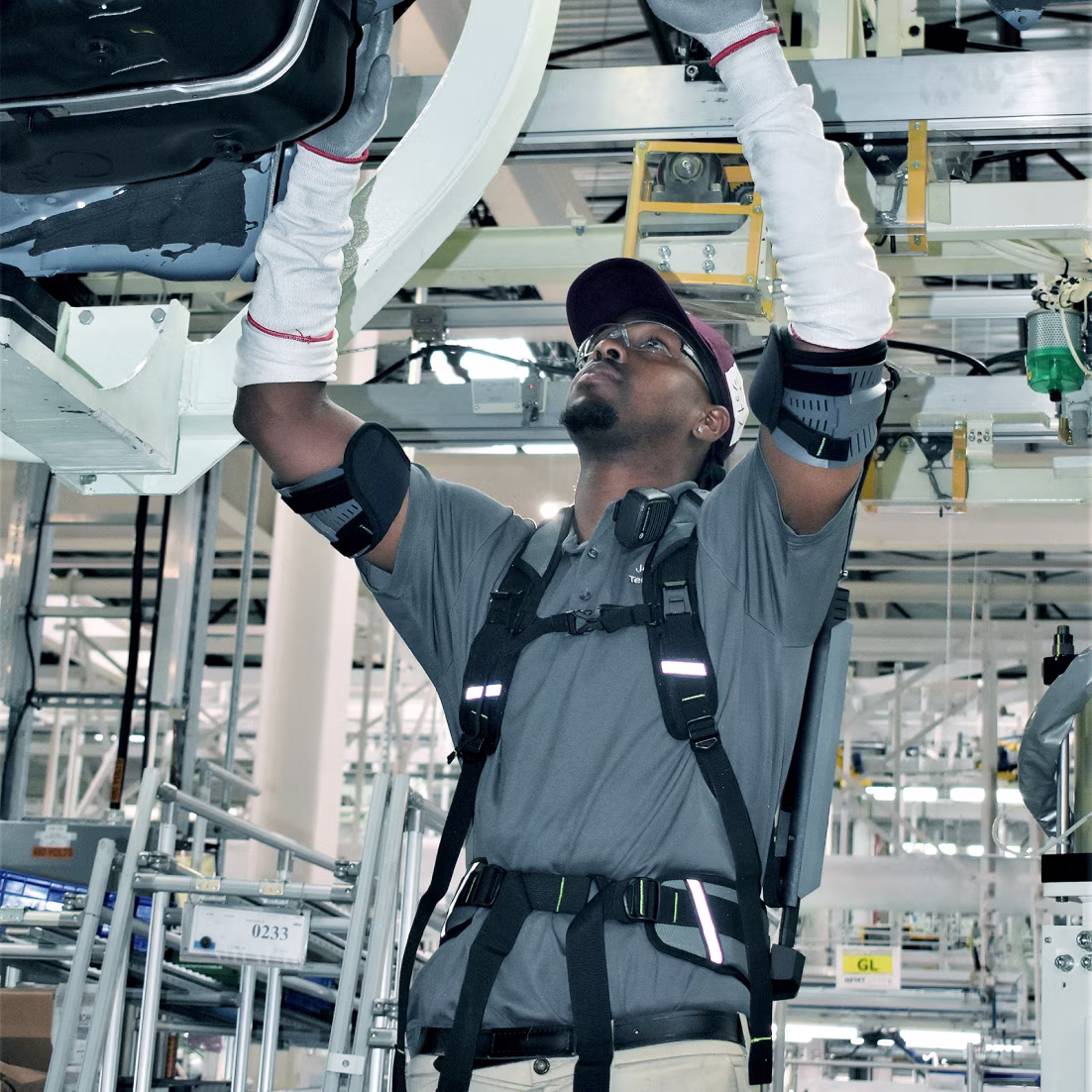 A worker with a shoulder exoskeleton stands in a factory hall under a vehicle and works overhead.