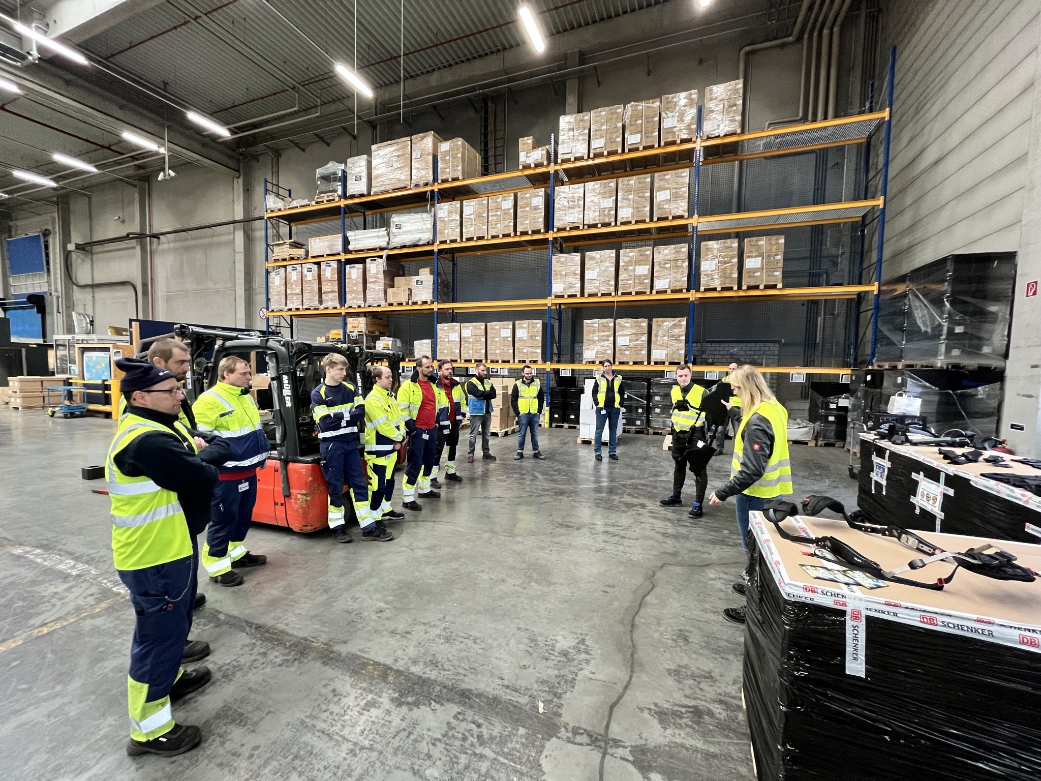 Zwei Mitarbeiter von SUITX by Ottobock unterweisen Lagerarbeiter bei DB Schenker in der Anwendung der Exoskelette. Im Hintergrund stehen Regale. Ein Arbeiter lehnt an einem Gabelstapler.