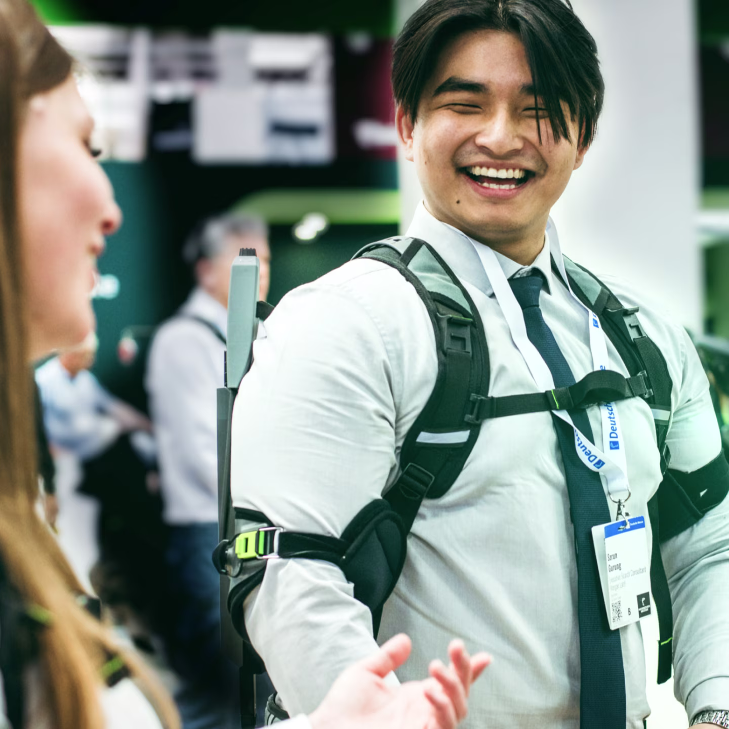 Mann auf trägt ein SUITX Exoskelett auf einer Messe und unterhält sich lächelnd mit einer Frau im Vordergrund 