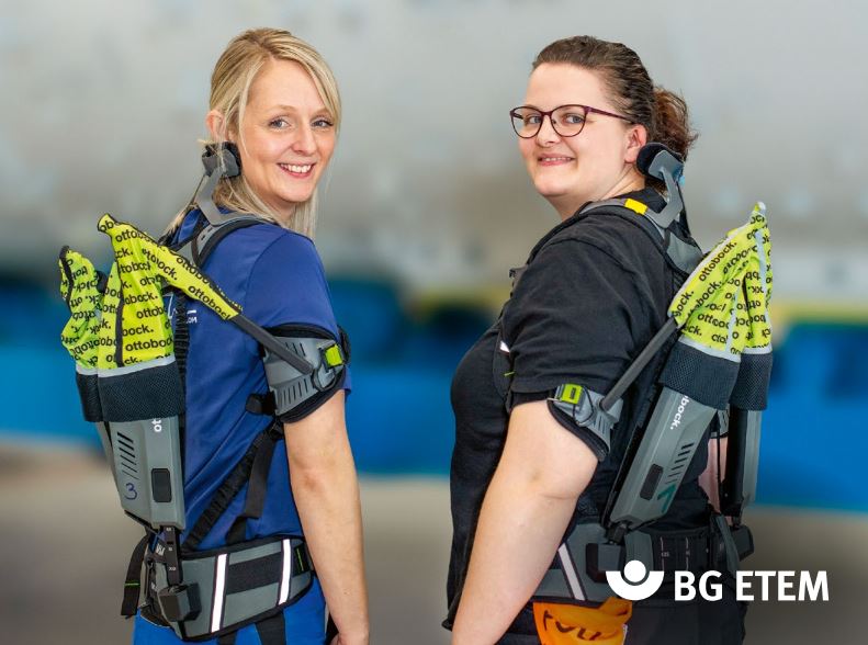 Two women wearing a shoulder exoskeleton look over their shoulders at the camera.