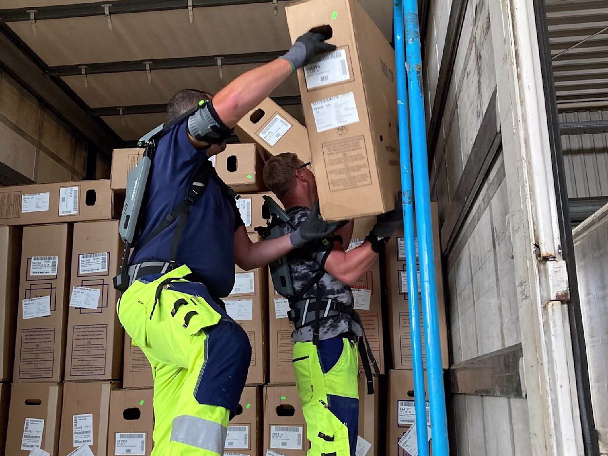 Two men wearing shoulder exoskeletons are unloading a truck.
