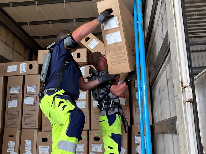 Two men wearing shoulder exoskeletons are unloading a truck.