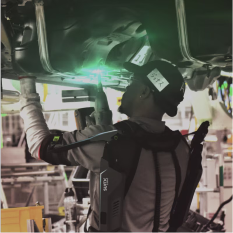 A male person in a factory during overhead assembly of a vehicle wears a SUITX IX SHOULDER exoskeleton