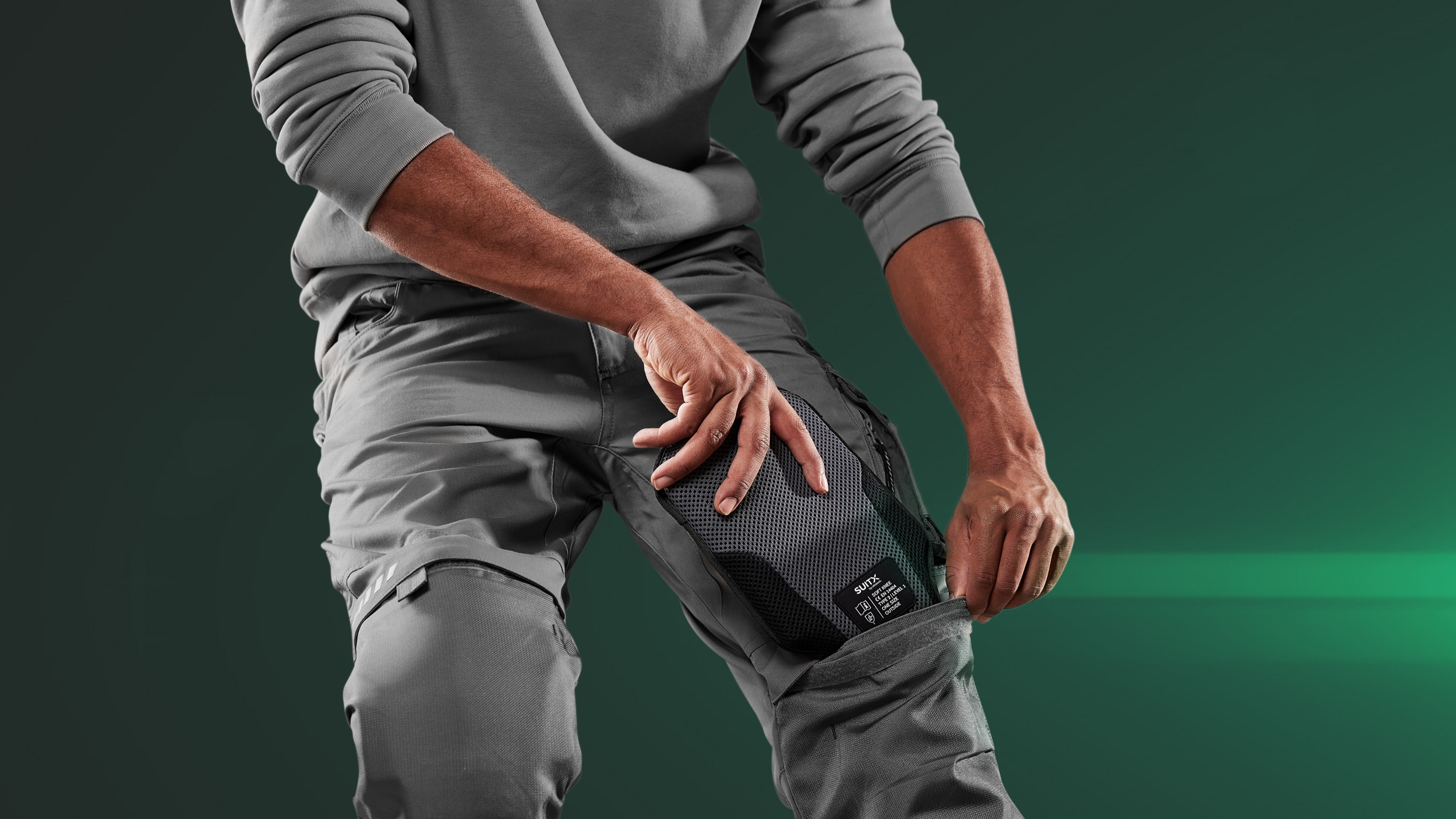 Partial view of a man in front of a dark green background putting the knee pad CX SOFT KNEE into the pocket of his work trousers.