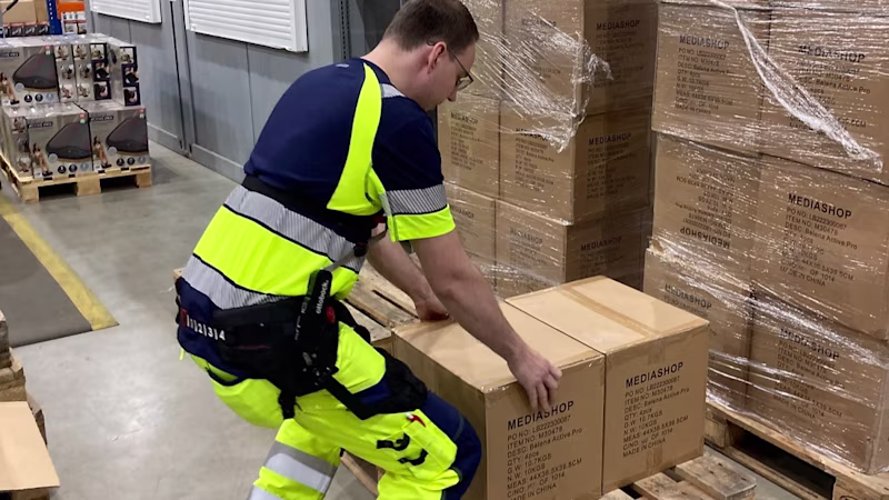A man wearing a back exoskeleton bends down to lift a crate from a pallet