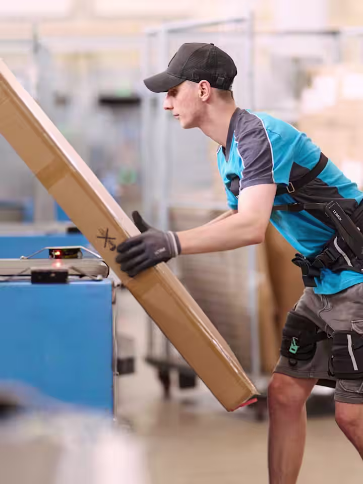 A warehouse worker in side view wears a back exoskeleton and is lifting a package onto a conveyor belt. (Photo: Hermes Germany / Willing-Holtz)