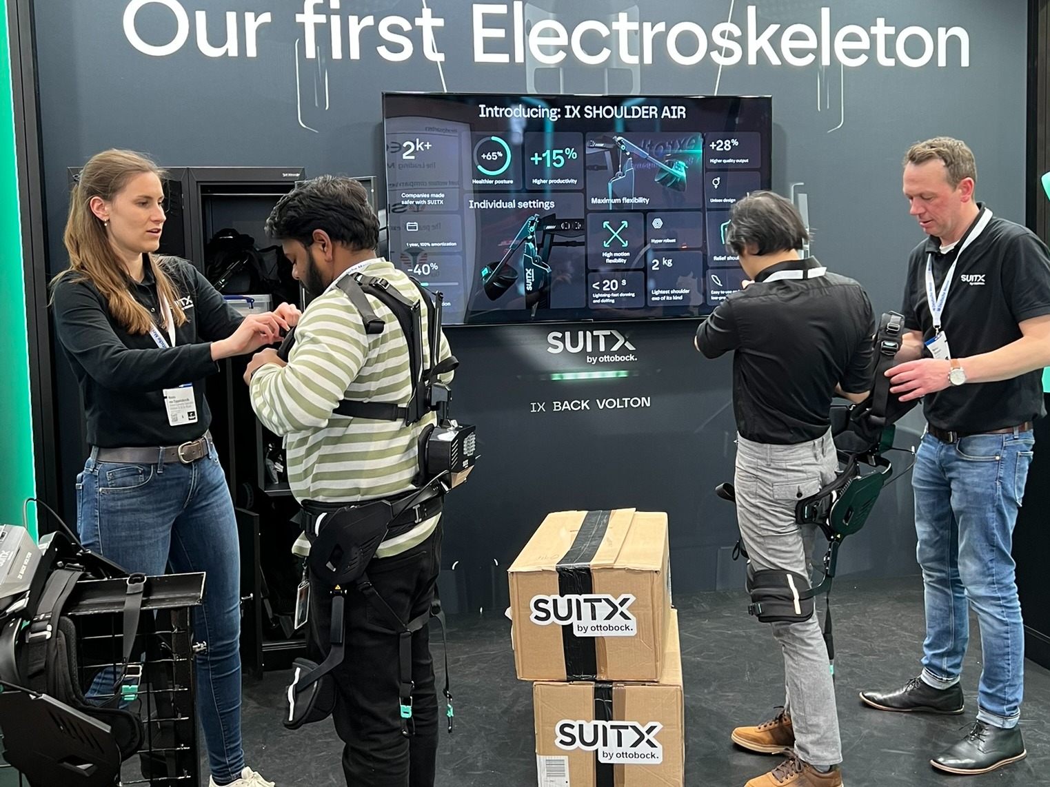 Two male visitors at a fair try on the IX BACK VOLTON exoskeleton suit while be supervised by two SUITX employees.
