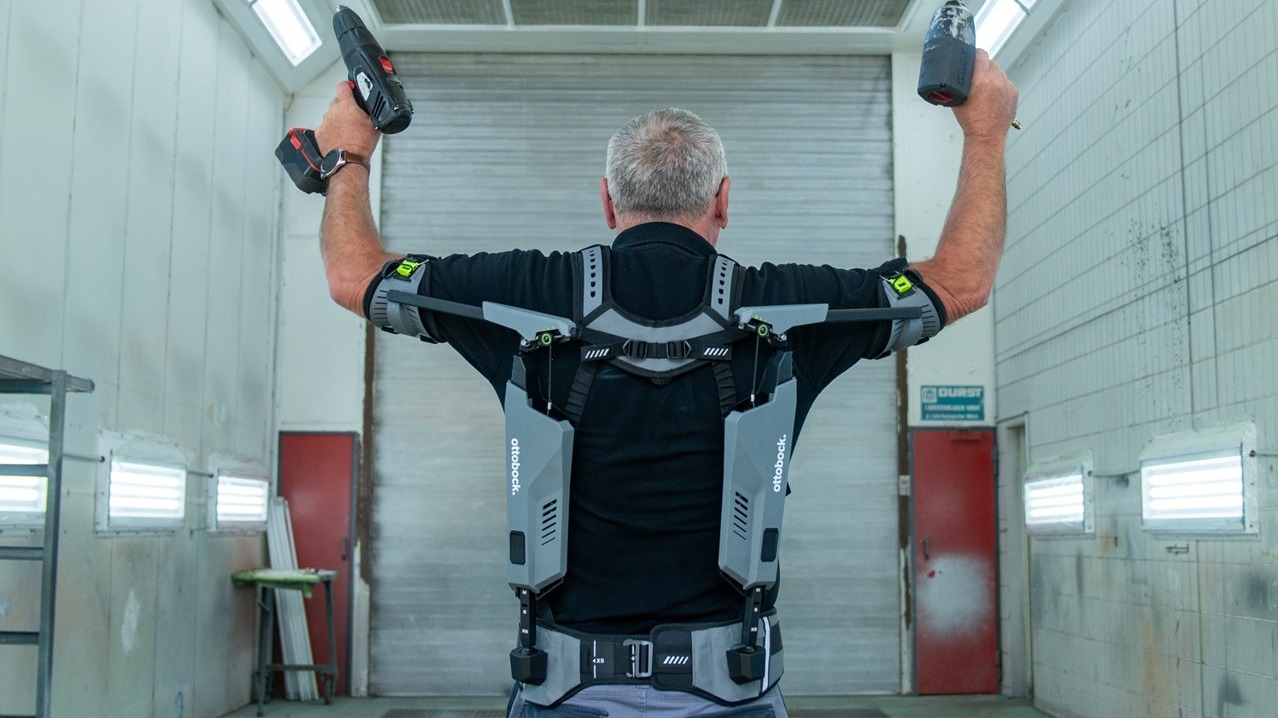 Back view of a mechanic wearing the IX SHOULDER shoulder exoskeleton and stretching both arms upwards. He is holding tools in his hands.