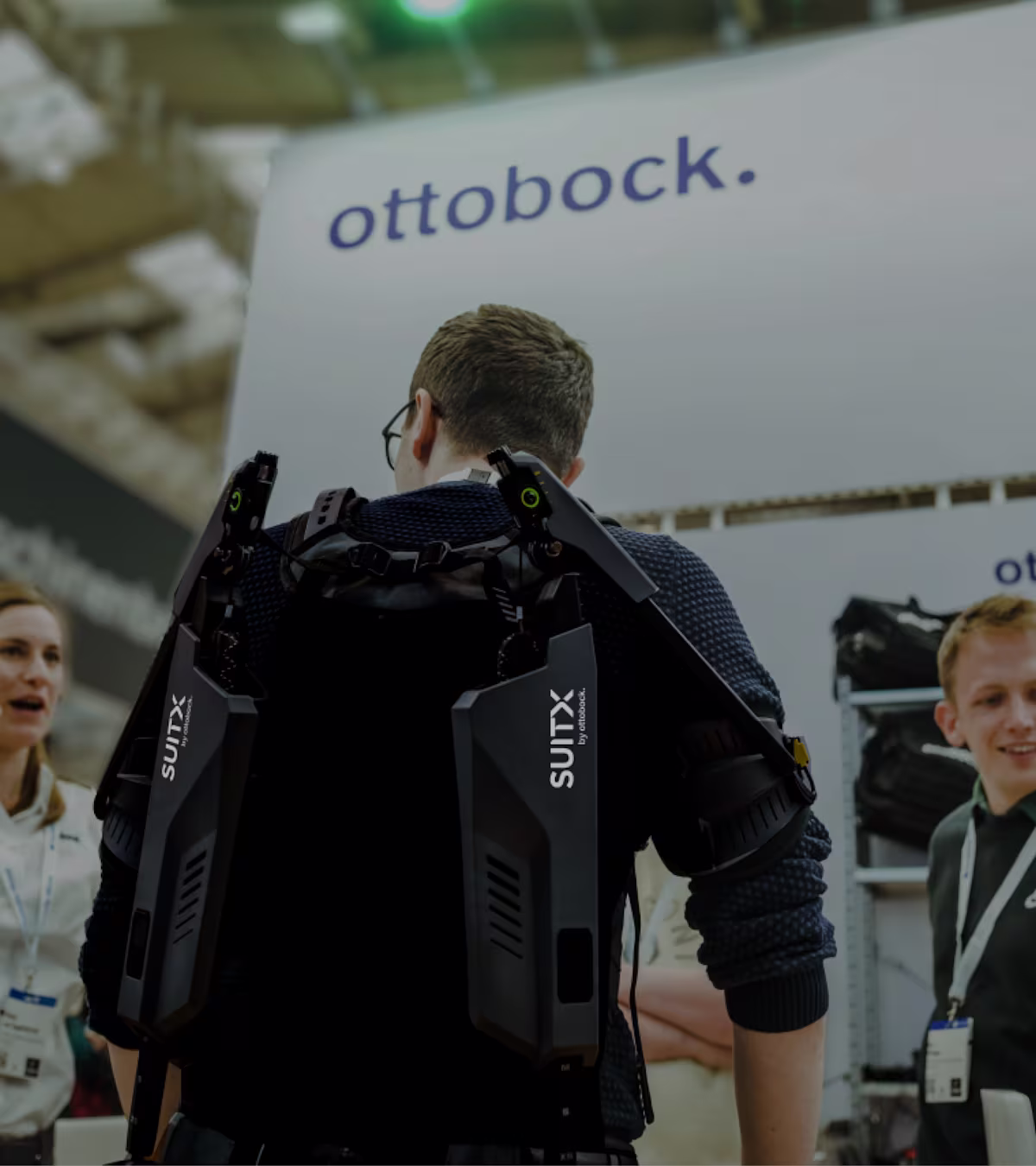Rear view of a male person wearing a SUITX exoskeleton at a trade show and talking to a woman to his left