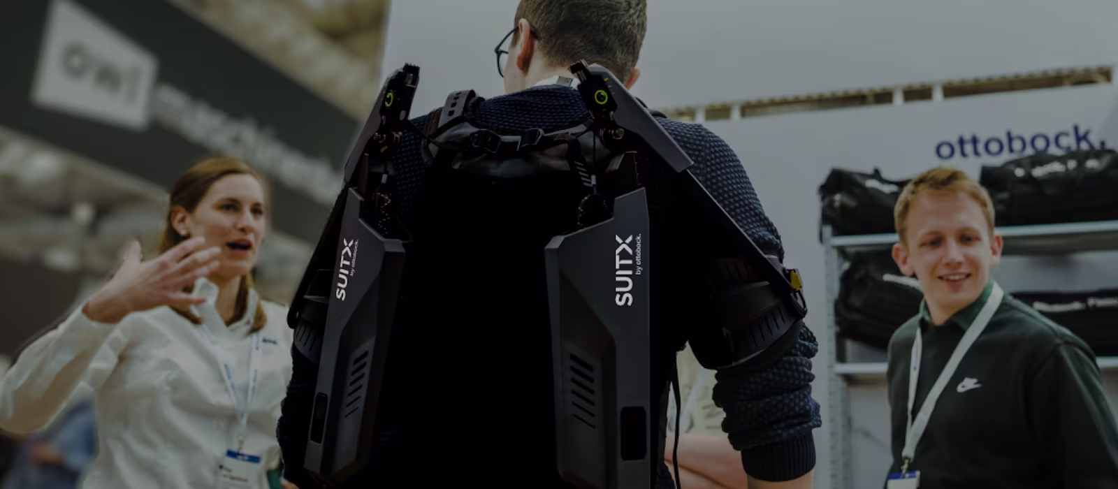 Rear view of a male person wearing a SUITX exoskeleton at a trade show and talking to a woman to his left