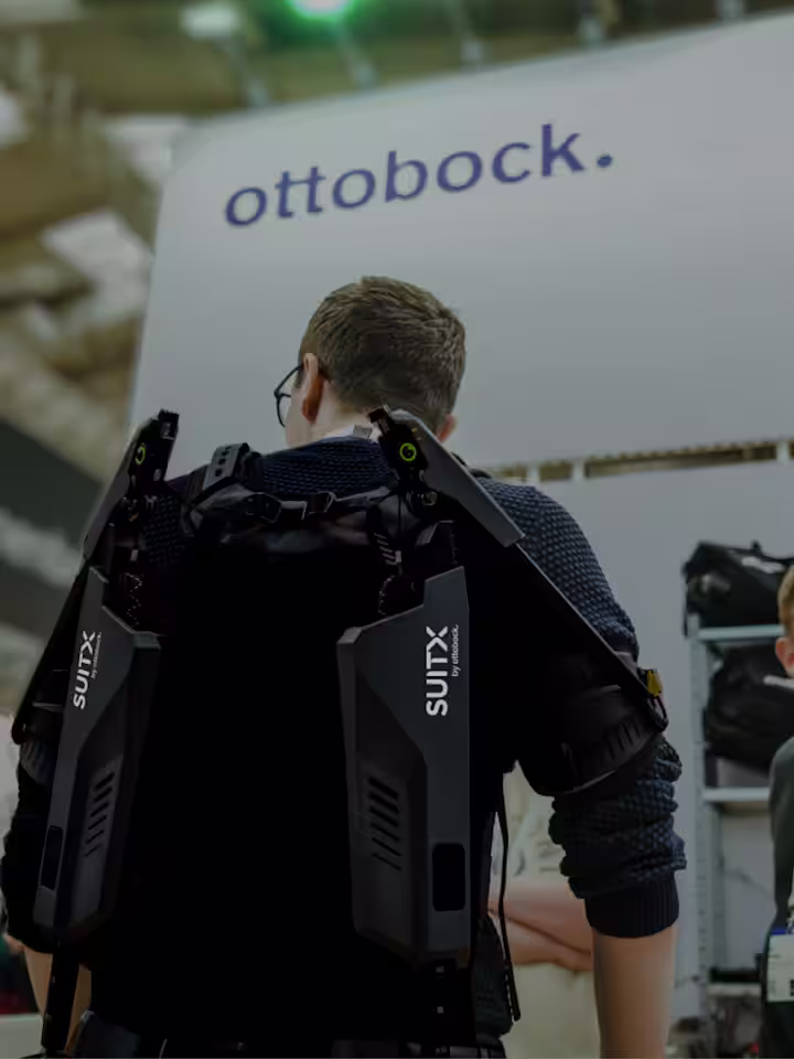 Rear view of a male person wearing a SUITX exoskeleton at a trade show and talking to a woman to his left