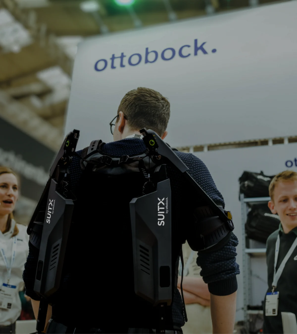 Rear view of a male person wearing a SUITX exoskeleton at a trade show and talking to a woman to his left