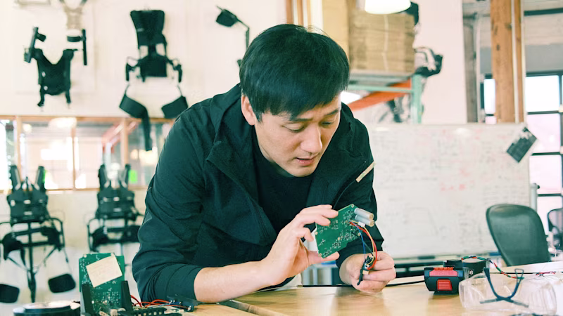 Developer Wayne leans against a work table and examines an electronic circuit board.