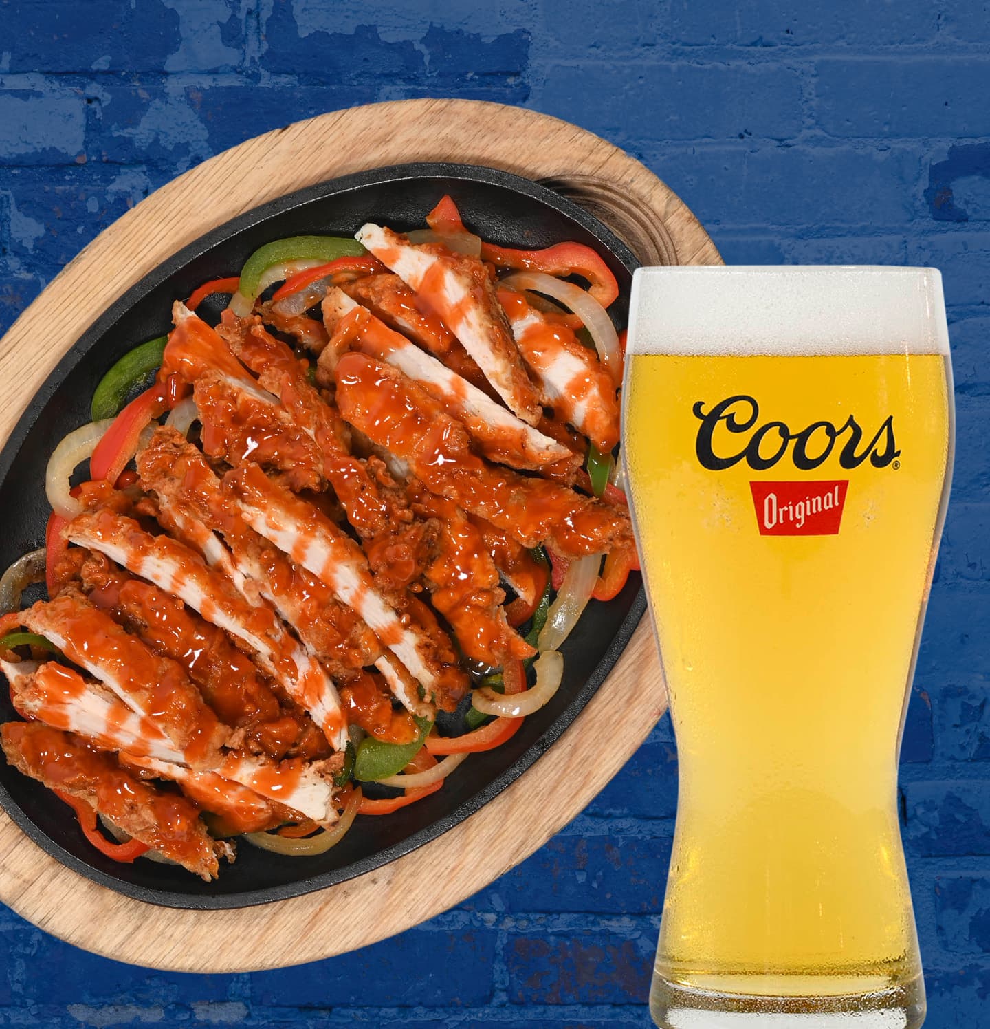 Sizzling skillet of buffalo-style chicken fajitas with grilled onions and bell peppers, served next to a pint of Coors Original beer on a wooden board and blue brick background.