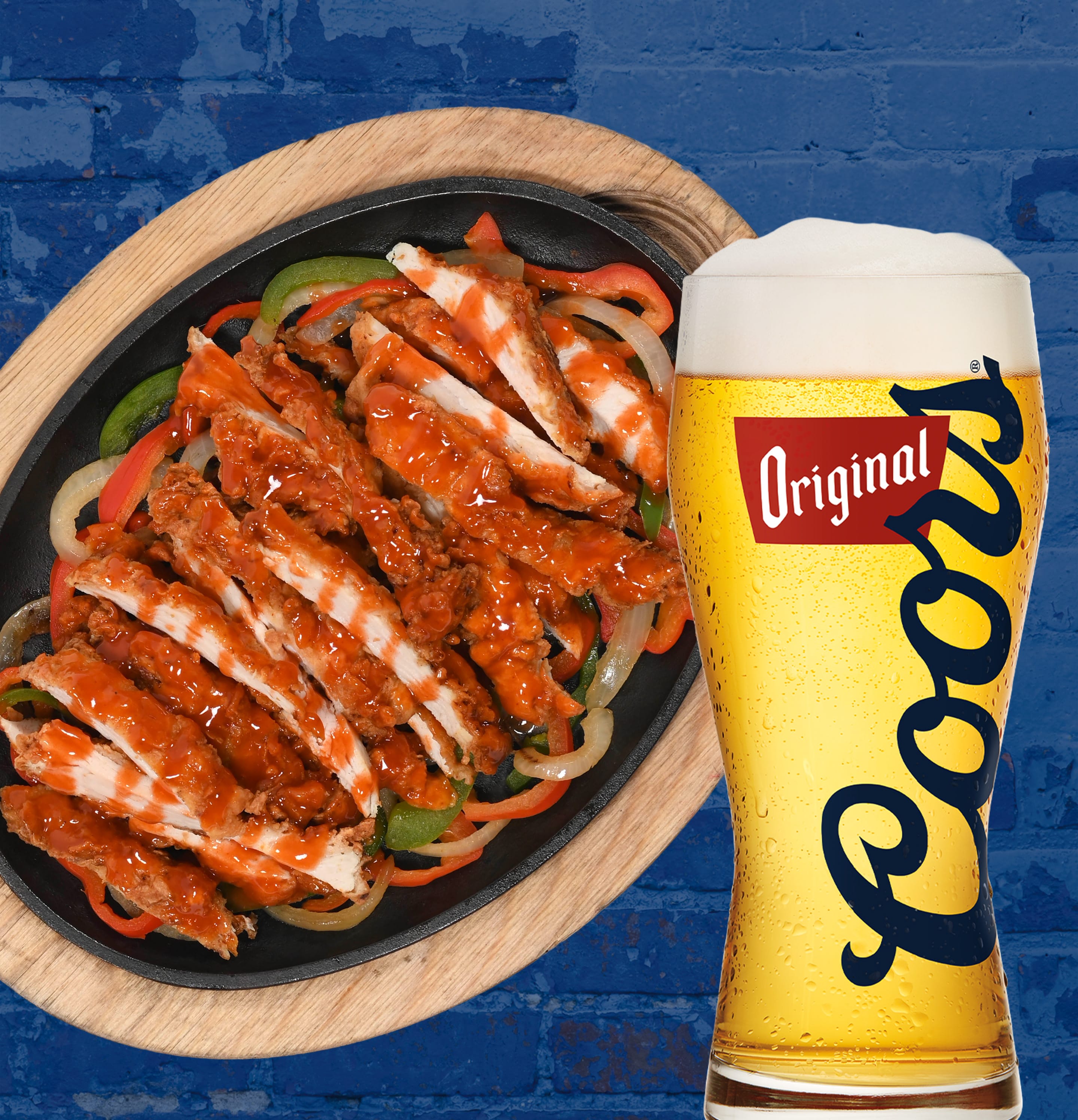 Sizzling skillet of buffalo-style chicken fajitas with grilled onions and bell peppers, served next to a pint of Coors Original beer on a wooden board and blue brick background.