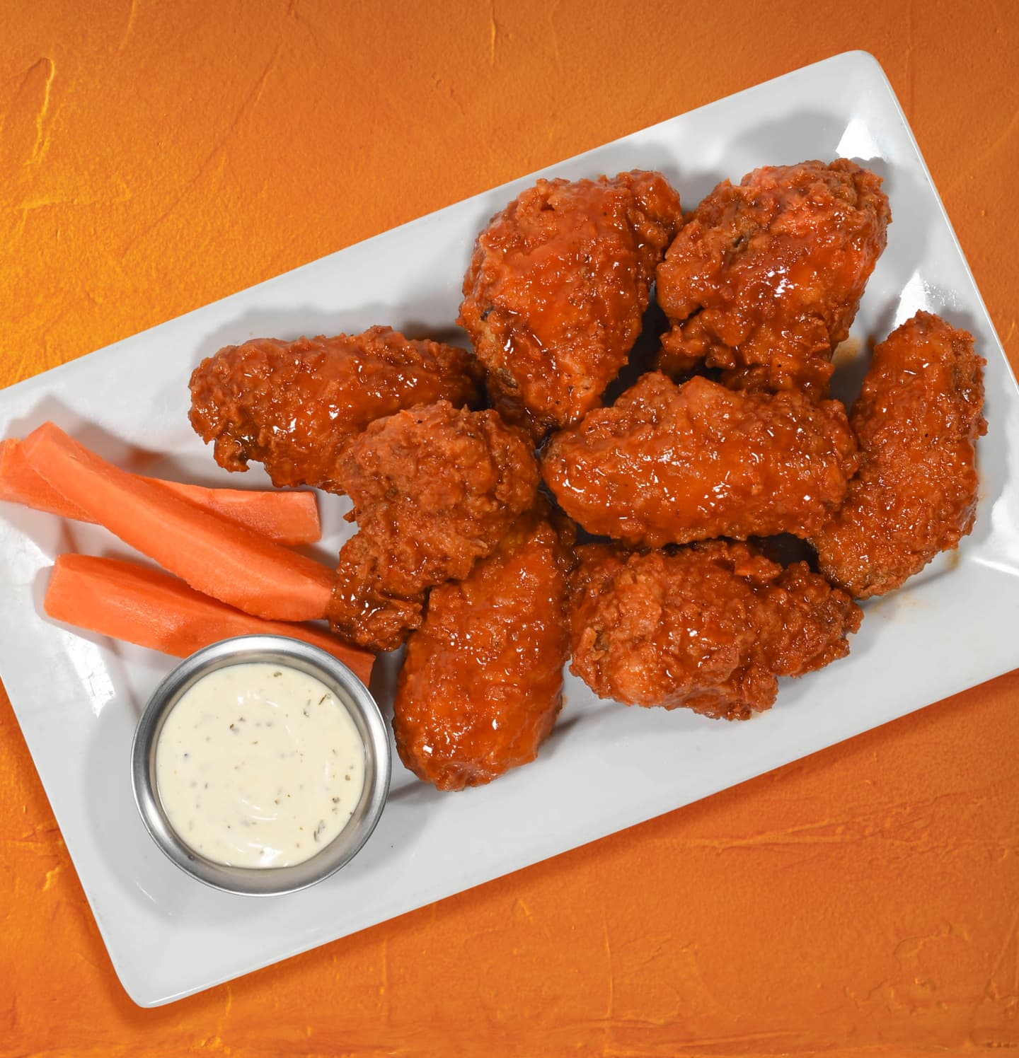 Plate of saucy buffalo chicken wings served with carrot sticks and a side of ranch dressing on a white plate against an orange background.