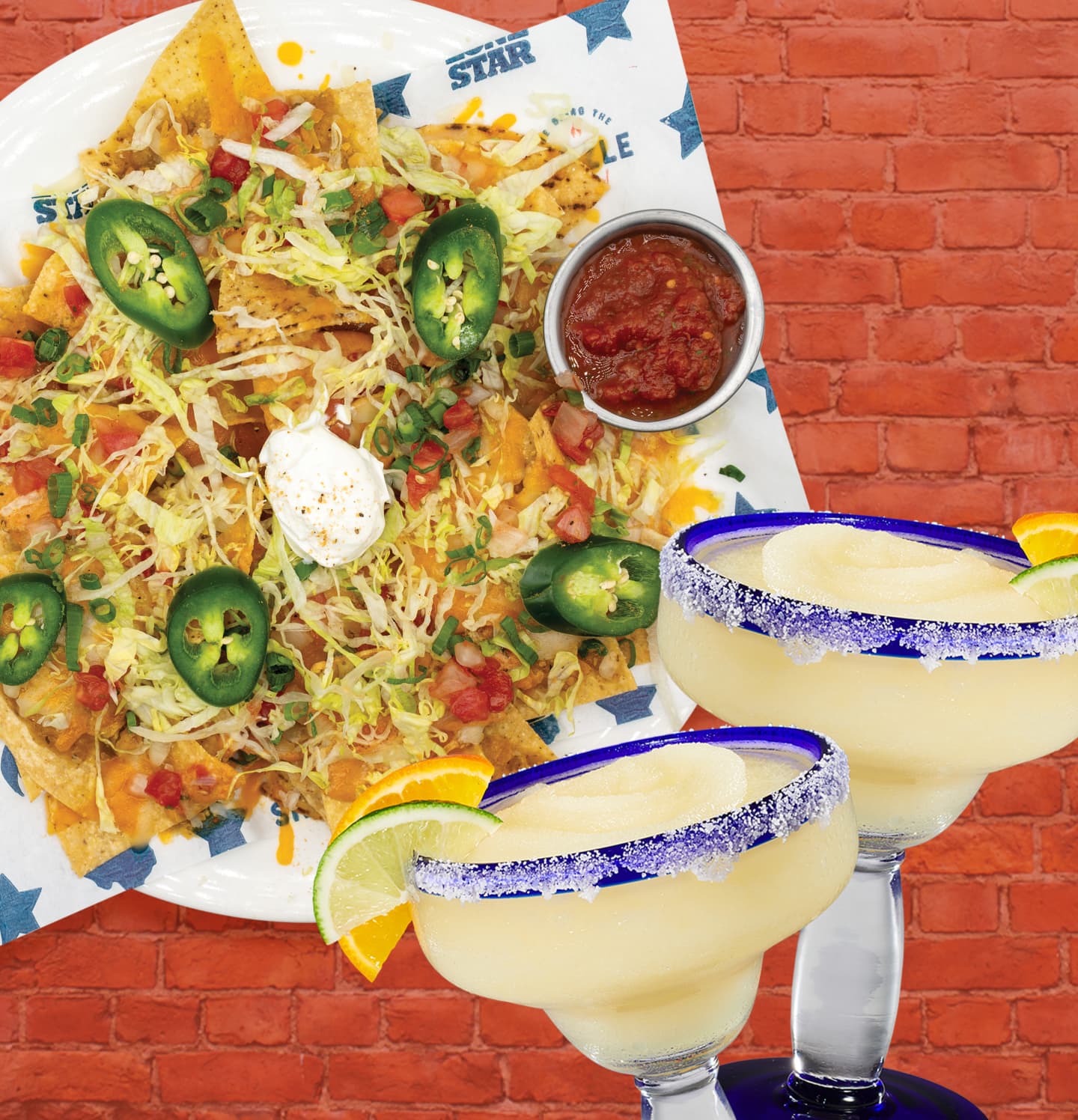 Plate of loaded nachos topped with lettuce, jalapeños, diced tomatoes, cheese, sour cream, and a side of salsa, served with two frozen lime margaritas against a red brick wall.