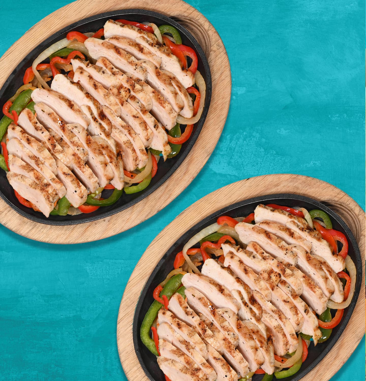 Two skillets of grilled chicken strips served over sautéed red and green bell peppers and onions, placed on a turquoise background.