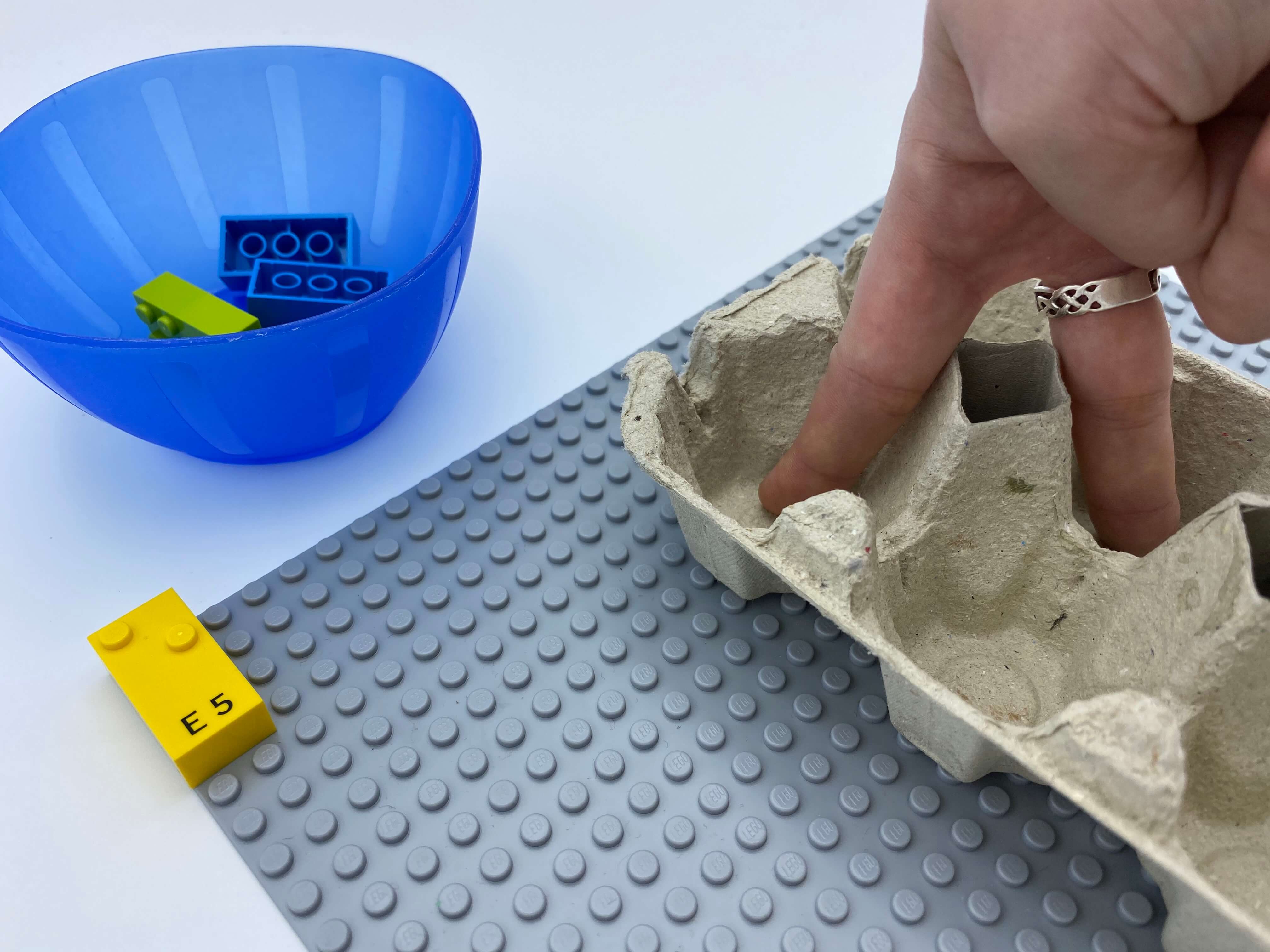 Letter brick e on the base plate, 2 fingers in an egg carton - 1 in dot 1 and 1 in dot 5, a bowl with bricks.