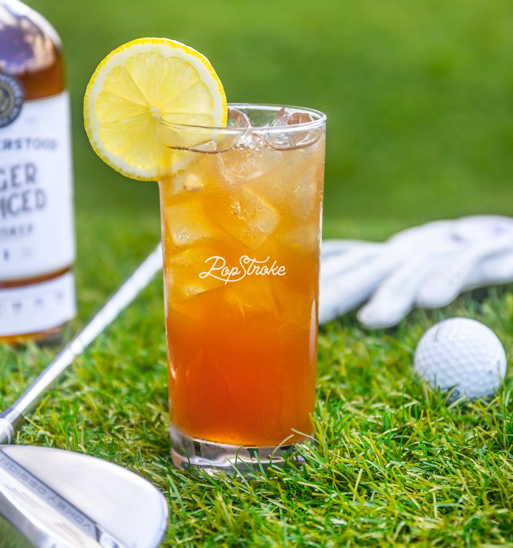 A glass holding an iced tea cocktail with a lemon slice in front of a golf club, set against a backdrop of green grass and a golf ball.
