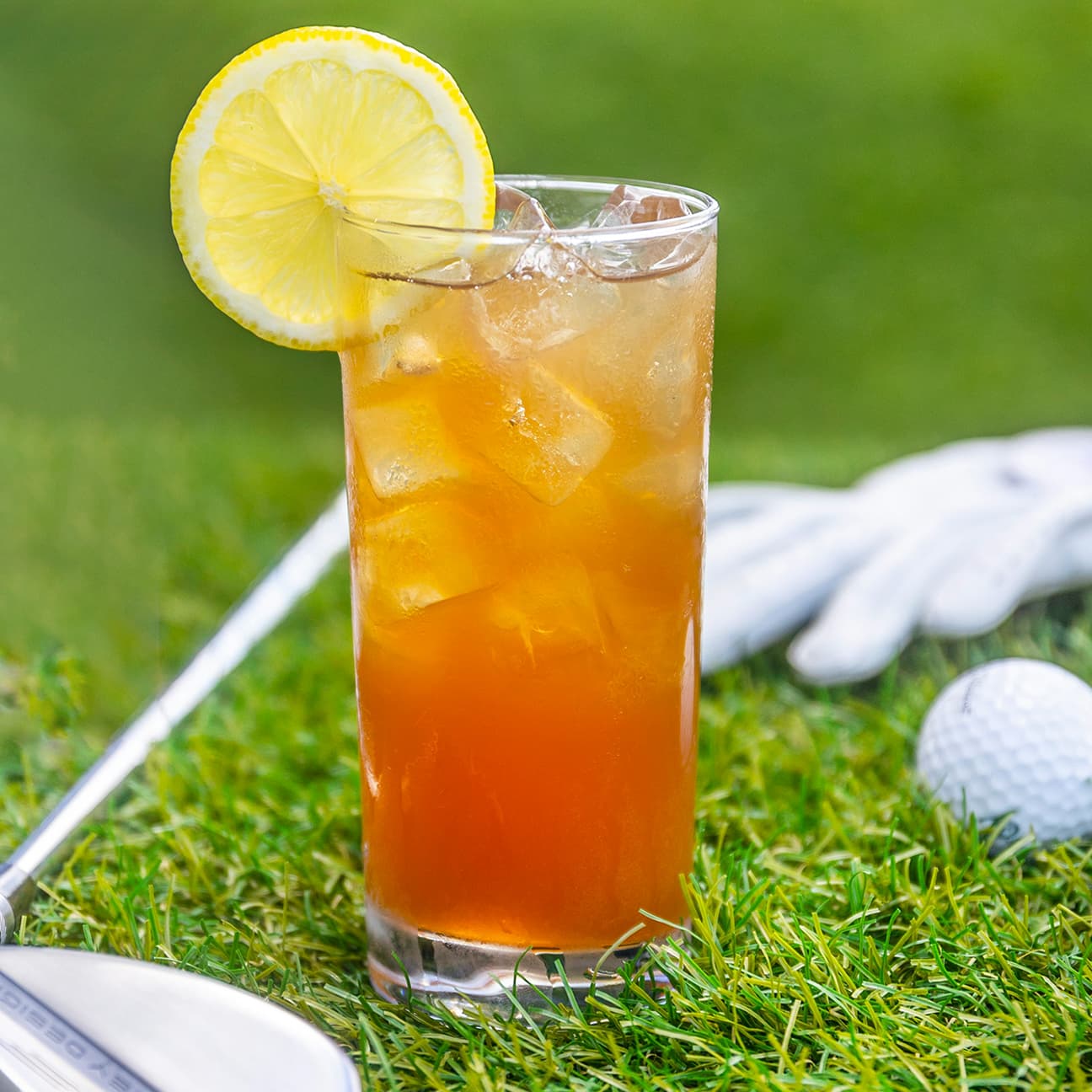 A glass holding an iced tea cocktail with a lemon slice in front of a golf club, set against a backdrop of green grass and a golf ball.