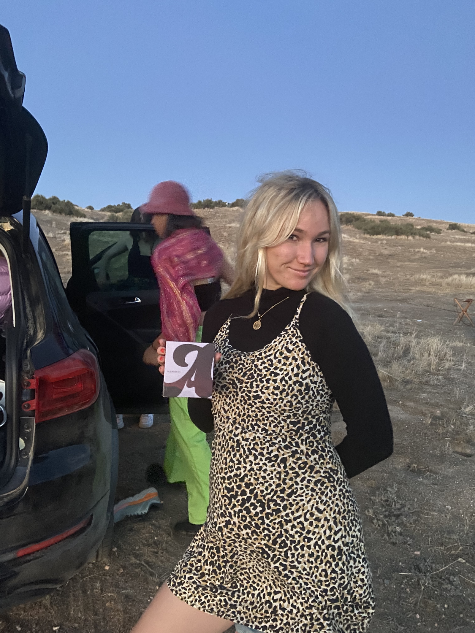 Person standing in the desert beside a car with open trunk and doors looking to the right and holding a box of august tampons