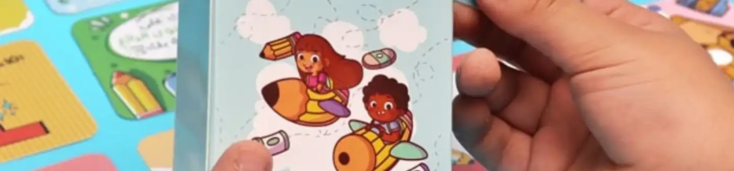 Hands holding a book with cartoon children riding pencils through clouds on the cover, set against colorful printed cards.